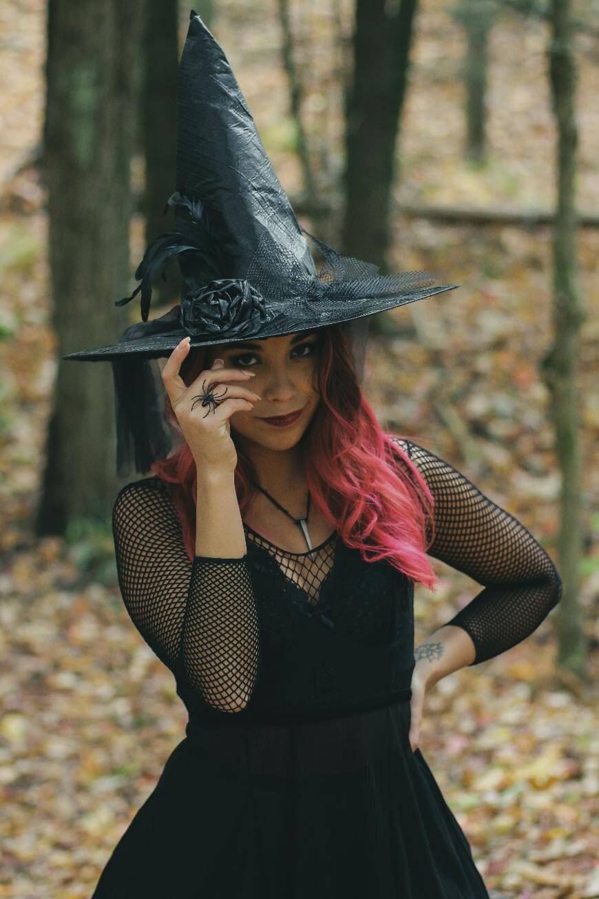 A person with pink hair, wearing a black witch hat and dress, stands in a forest. They are holding a fake spider near their face and smiling mischievously. The background is filled with trees and fallen leaves.