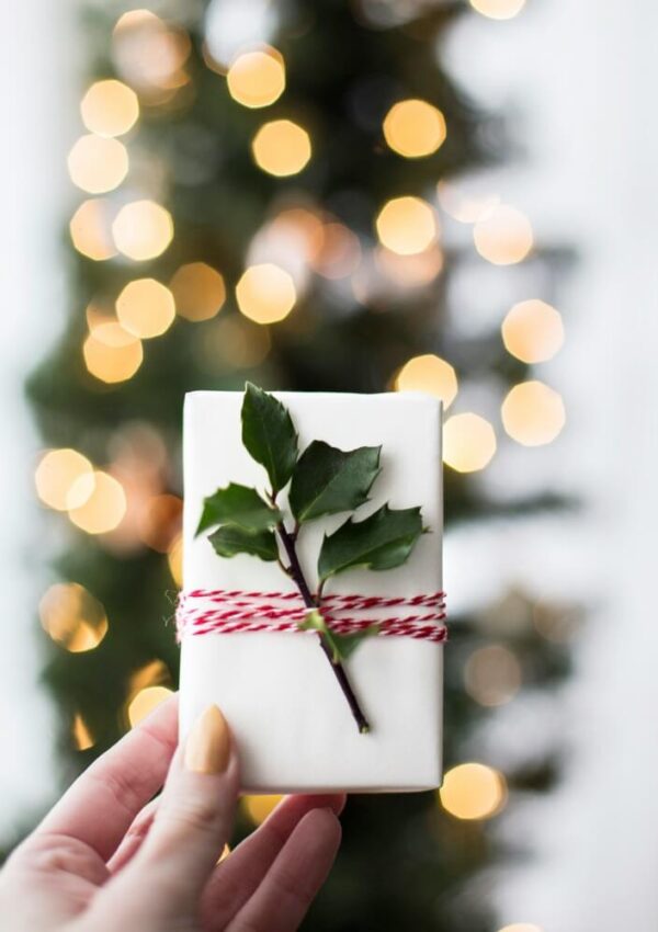 A hand holds a small, white gift wrapped with red and white twine and decorated with a sprig of holly; a Christmas tree with glowing lights is blurred in the background.
