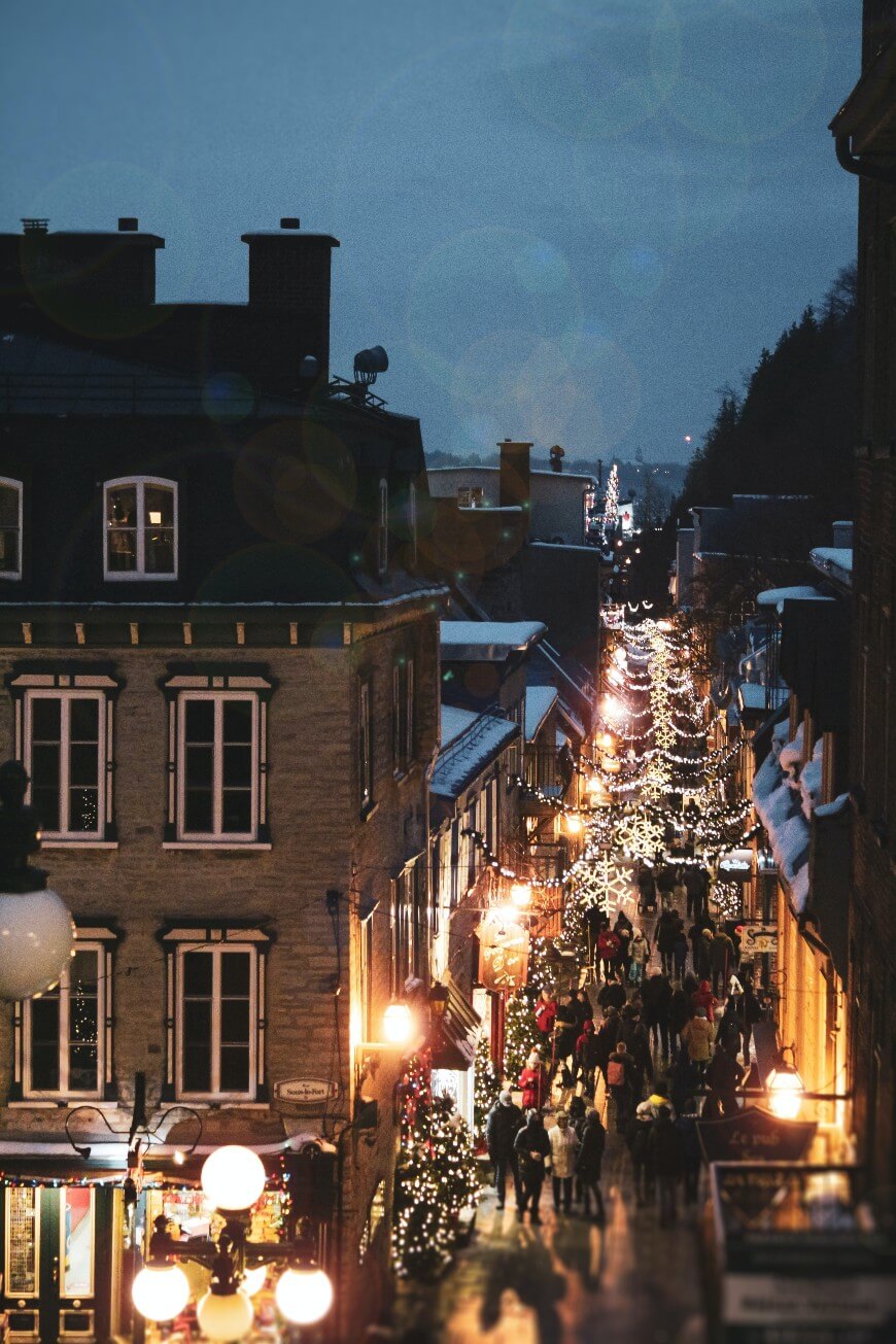 A bustling street at dusk, lined with old buildings and decorated with festive lights. Crowds of people walk below strings of glowing lights, creating a warm, lively atmosphere in the evening.