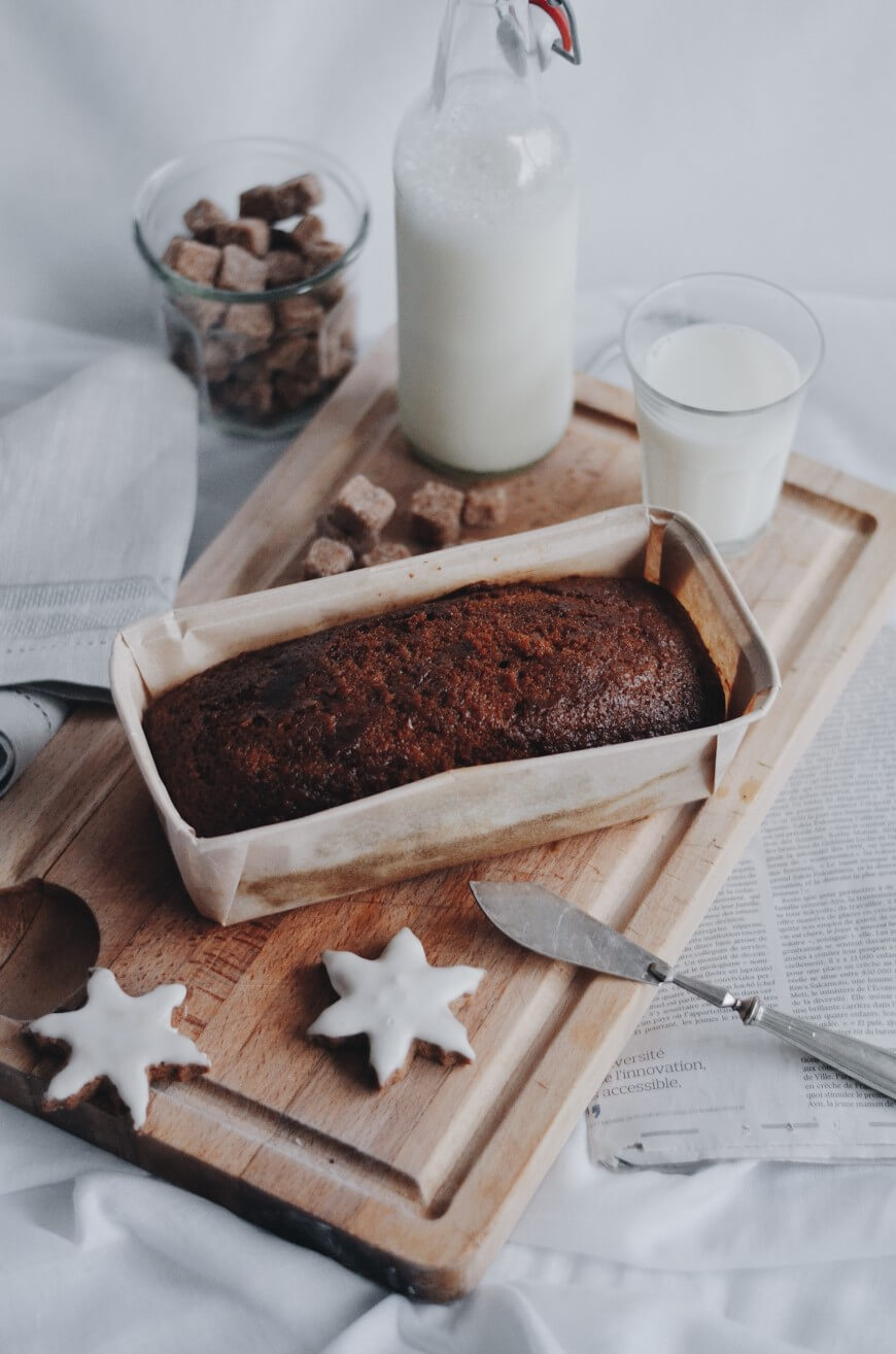 A loaf of dark bread in parchment paper on a wooden board, next to a glass and bottle of milk, sugar cubes, a butter knife, and two star-shaped iced cookies.
