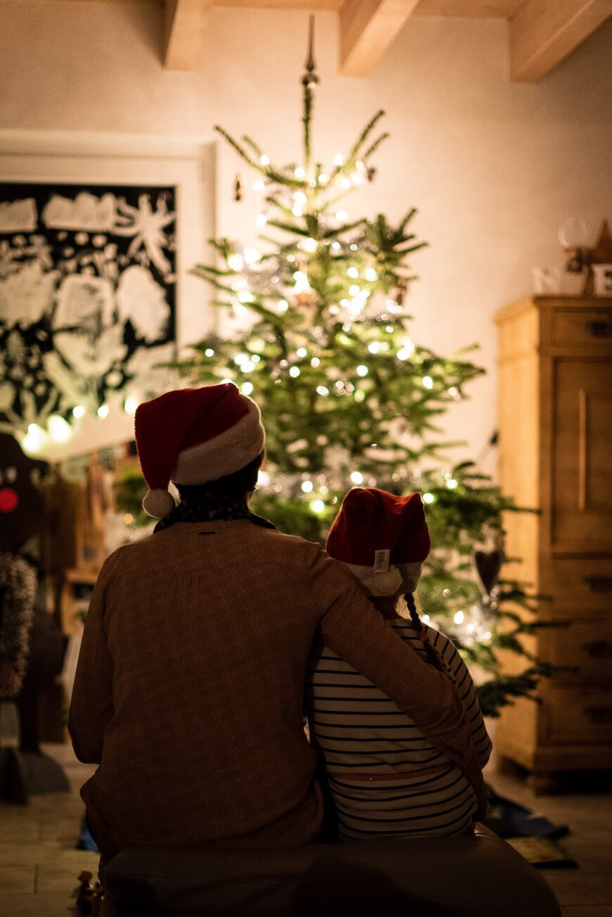 Two people wearing Santa hats sit close together, facing a decorated Christmas tree with glowing lights in a cozy, warmly lit room. One has their arm around the other, creating a festive and affectionate atmosphere.