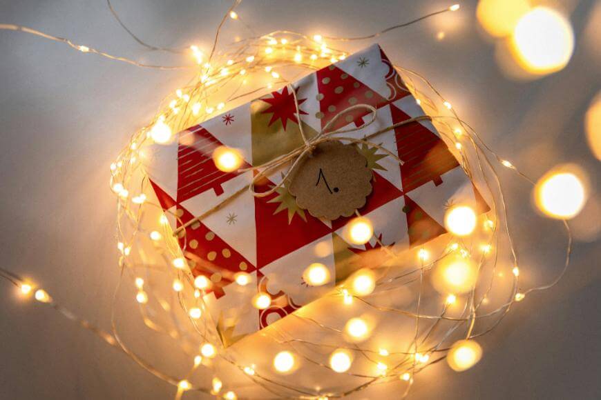 A festively wrapped gift with red and gold patterns is surrounded by glowing string lights. A brown tag with the number “1” tied to the gift suggests it is part of an advent calendar or countdown.