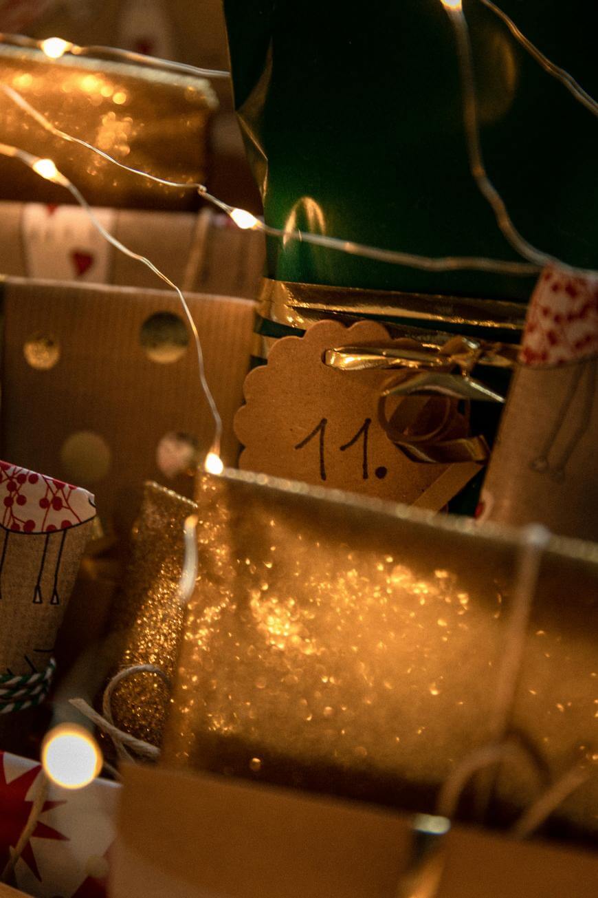A close-up of several wrapped gifts with gold glittery paper and brown tags, one labeled 11. Warm fairy lights are draped over the presents, creating a festive and cozy atmosphere.