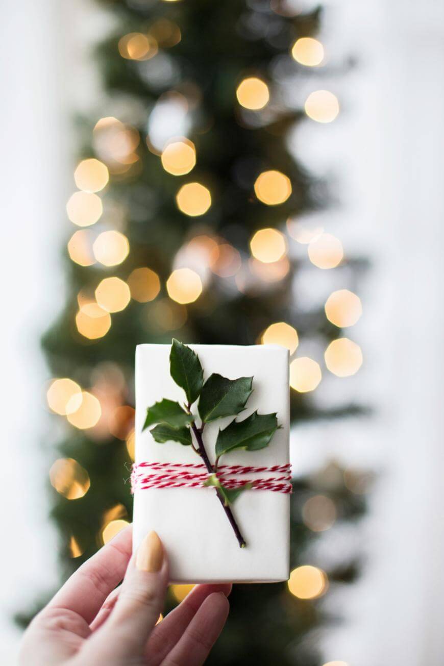A hand holds a small white gift wrapped with red and white string and decorated with a sprig of holly; blurred Christmas tree lights glow warmly in the background.
