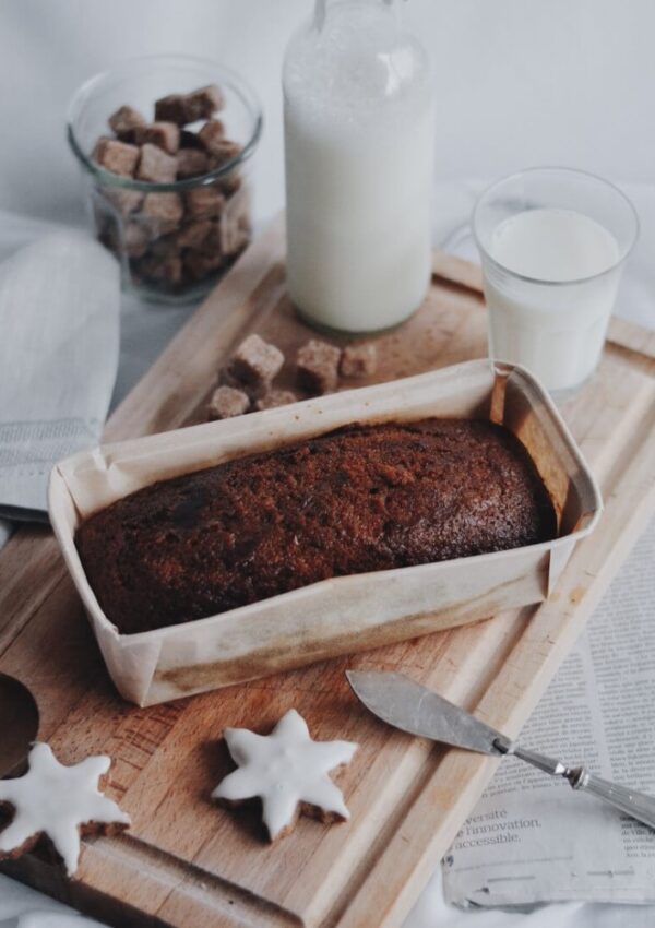 A loaf of banana bread in parchment on a wooden board, next to a knife, two star-shaped iced cookies, a glass and bottle of milk, a bowl of brown sugar cubes, and a folded newspaper.