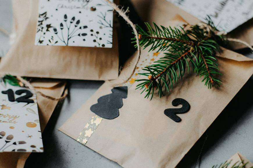Brown paper gift bags decorated with twine, evergreens, numbered tags, and festive cards with holiday patterns, arranged on a surface for an advent calendar or Christmas celebration.