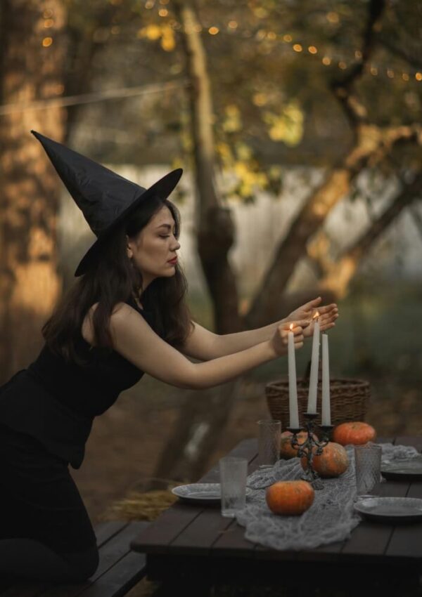 A woman in a black witch hat and dress kneels by an outdoor table, lighting white candles. The table is decorated with small pumpkins, glassware, and a gray cloth. Autumn trees and soft lights are in the background.