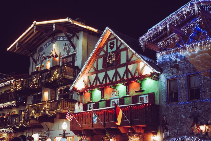 Festively decorated buildings with alpine architecture are lit up at night with colorful lights, garlands, and flags, evoking a charming winter village atmosphere.