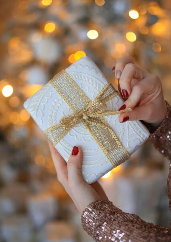 A person with red nail polish holds a white gift wrapped with gold ribbon, in front of a blurred, festive background with warm lights. The person wears a sparkly, sequined sleeve.