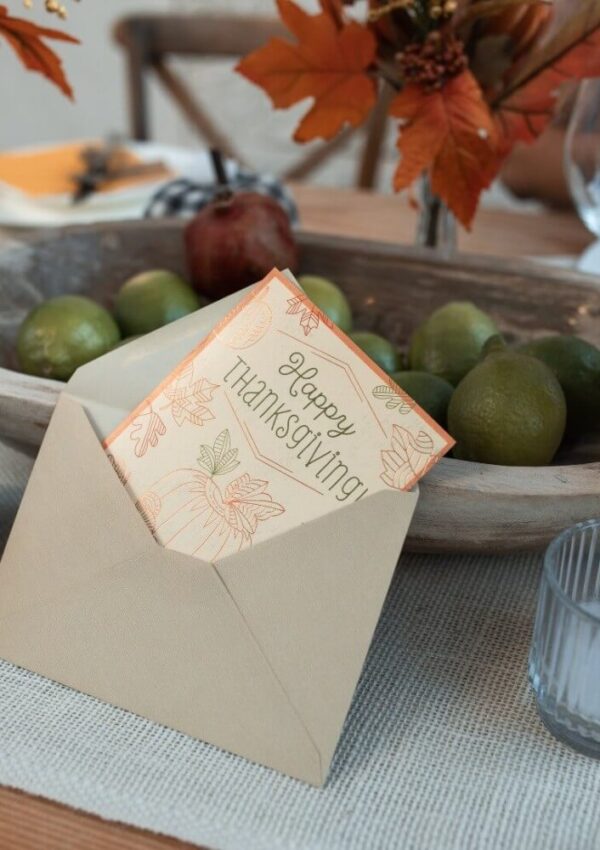 A Thanksgiving card in an open beige envelope sits on a table near a wooden bowl of green limes and autumn leaves, with a glass and candle nearby.
