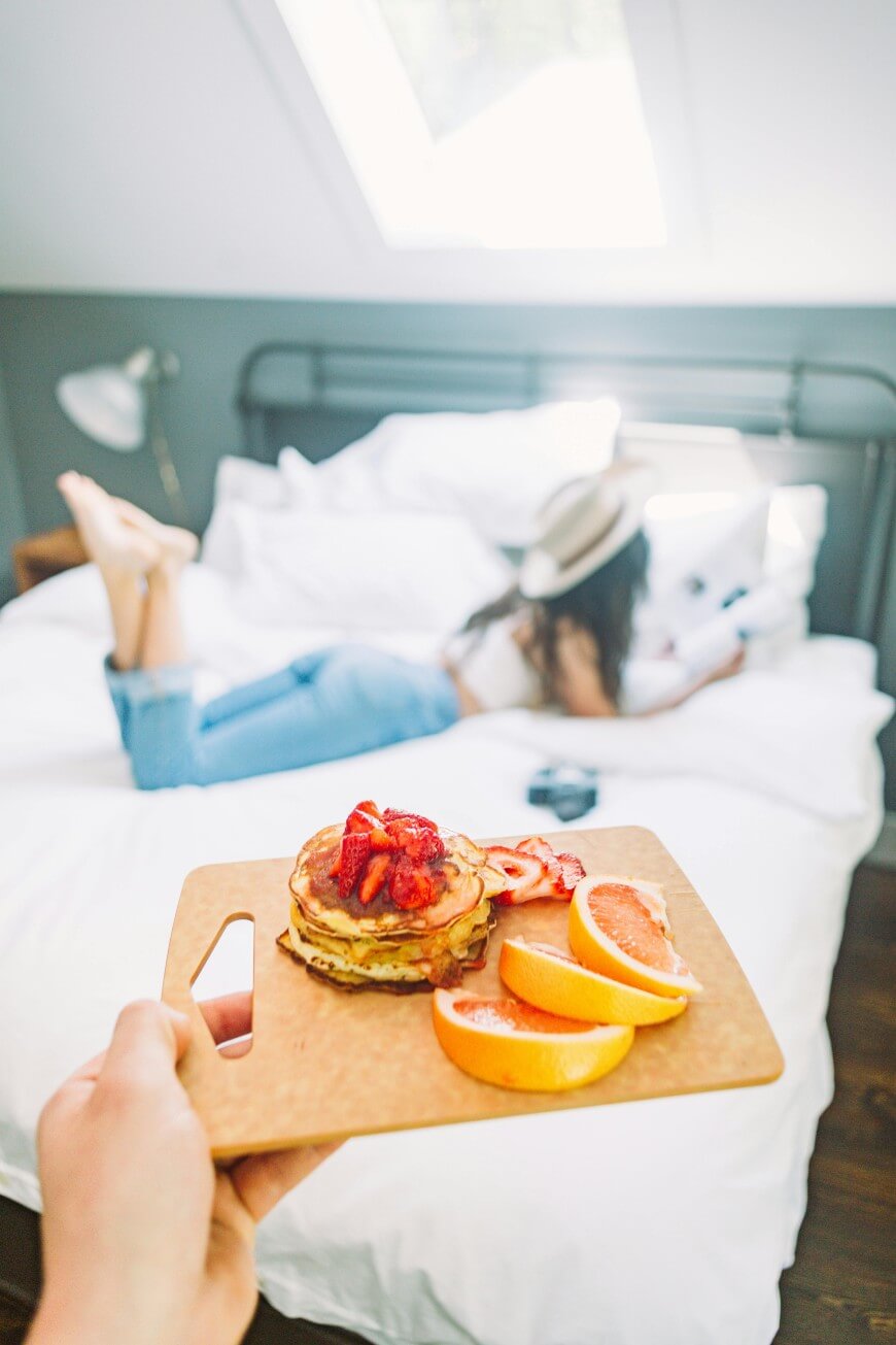 A person holds a wooden board with pancakes topped with strawberries and orange slices in the foreground; in the background, a woman in jeans and a hat reads on a white bed under a skylight.