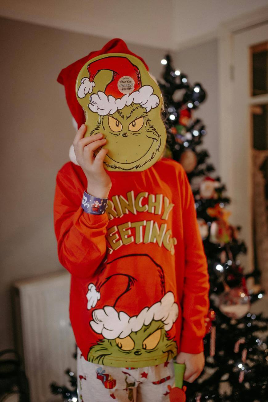 A child wearing a red Grinchy Greetings shirt and pajama pants stands by a decorated Christmas tree, holding a Grinch mask in front of their face, playfully concealing their identity.