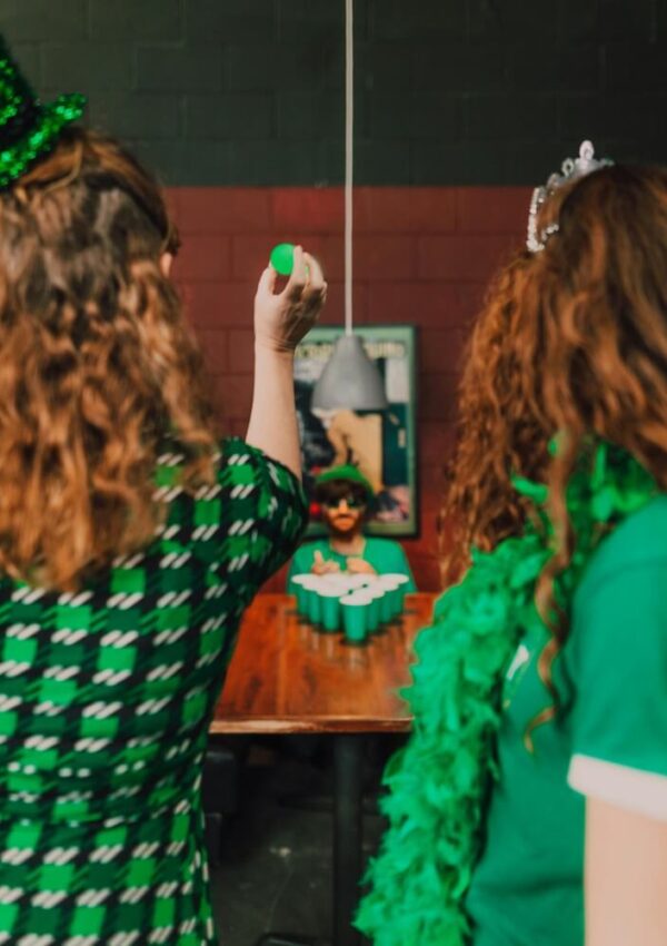 Two people with curly hair wearing green St. Patrick’s Day-themed outfits play beer pong, aiming a ball at cups on a table. The background includes a poster and a hanging light.