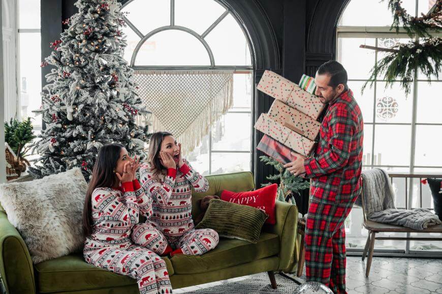 A man in plaid pajamas holds a tall stack of Christmas gifts, surprising two women in matching holiday pajamas who are sitting excitedly on a green couch near a decorated Christmas tree.