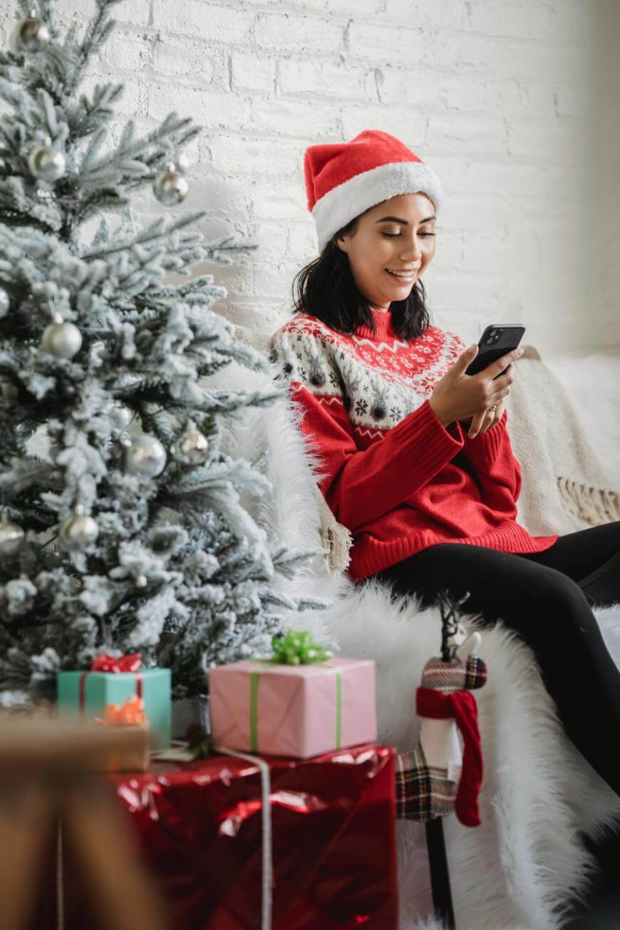 A woman in a Santa hat and festive sweater sits by a decorated Christmas tree, smiling while looking at her phone. Wrapped gifts with ribbons are placed under the tree.