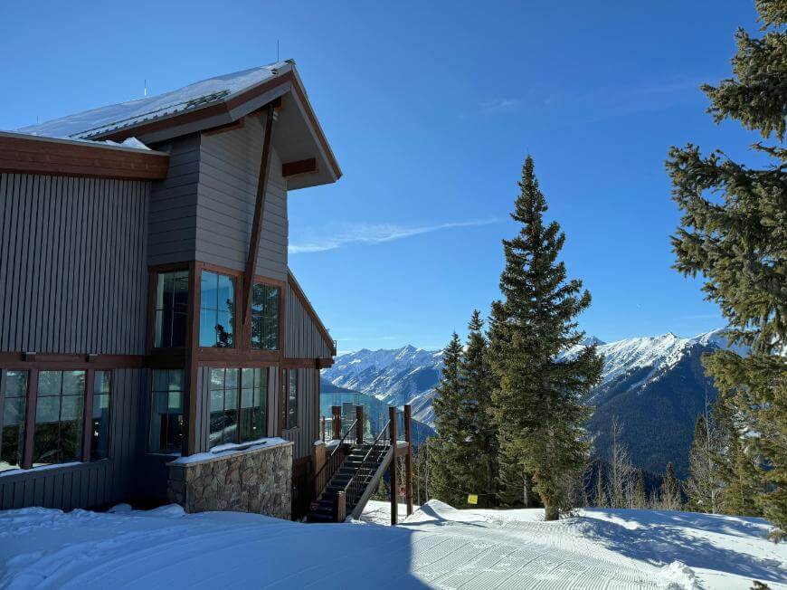 A modern mountain lodge with large windows sits on a snowy slope, surrounded by pine trees, with snow-covered mountains and a clear blue sky in the background.