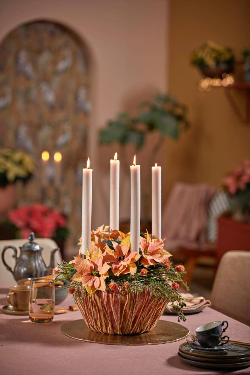 A table centerpiece with four tall lit candles surrounded by peach and pink flowers in a brown vase, set on a pink tablecloth with teacups, saucers, and a teapot in a cozy, softly lit room.