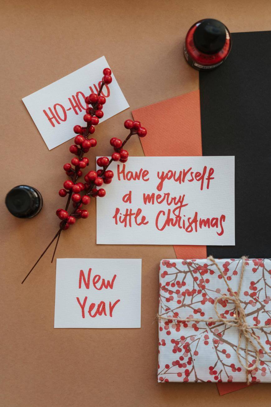 A flat lay of holiday cards with red handwritten messages, a sprig of red berries, black ink bottles, and a gift wrapped in berry-patterned paper with twine, on a brown background. Cards read “HO-HO-HO,” “New Year,” and “Have yourself a merry little Christmas.”.