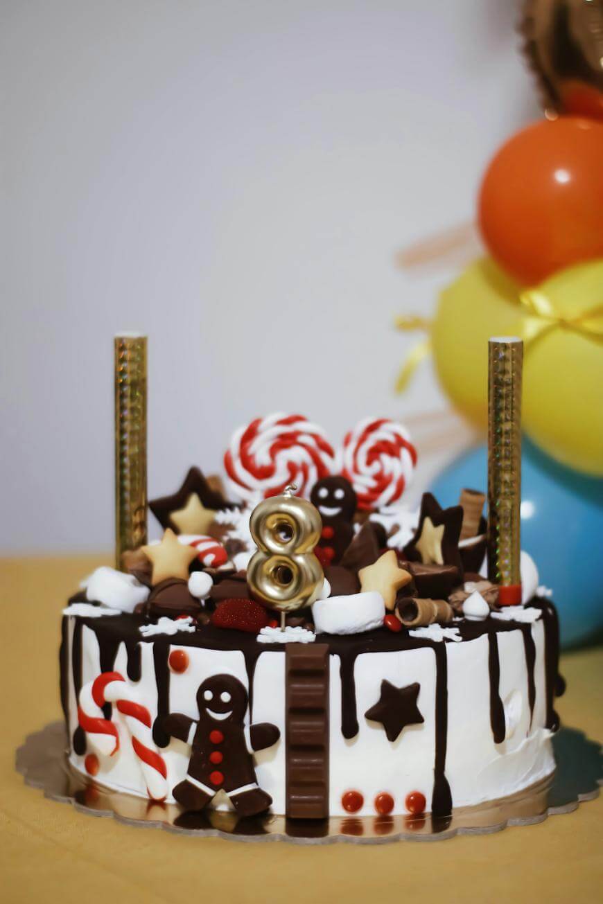 A decorated birthday cake with chocolate drizzle, gingerbread men, candy canes, lollipops, and a gold number 8 candle. Colorful balloons are in the background.