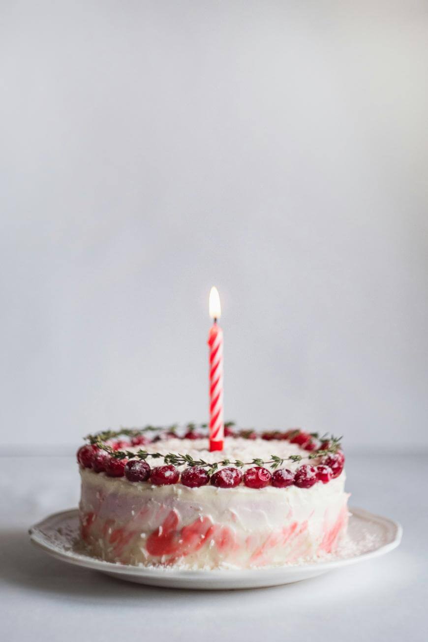 A round cake with white and pink marbled frosting, topped with red berries and a ring of green herbs, sits on a white plate. A single lit, red-and-white striped candle is placed in the center.