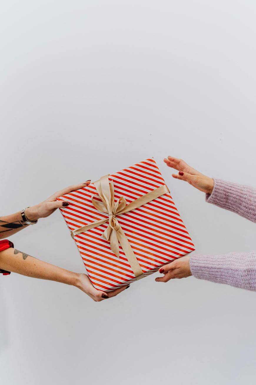 Two people exchange a large, square, red-and-white striped gift box with a gold ribbon and bow against a plain white background. Only their hands and part of their arms are visible.