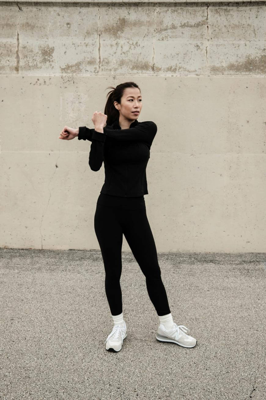 A woman in black athletic wear and white sneakers stretches her arm across her chest while standing on a paved surface in front of a concrete wall.