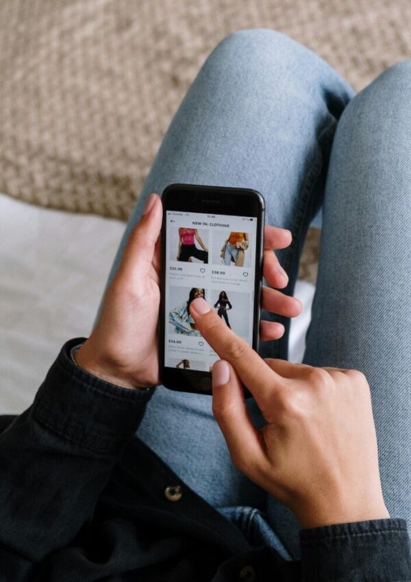 A person sitting cross-legged on a bed shops online using a smartphone, scrolling through a clothing store app that displays images and prices of various outfits.