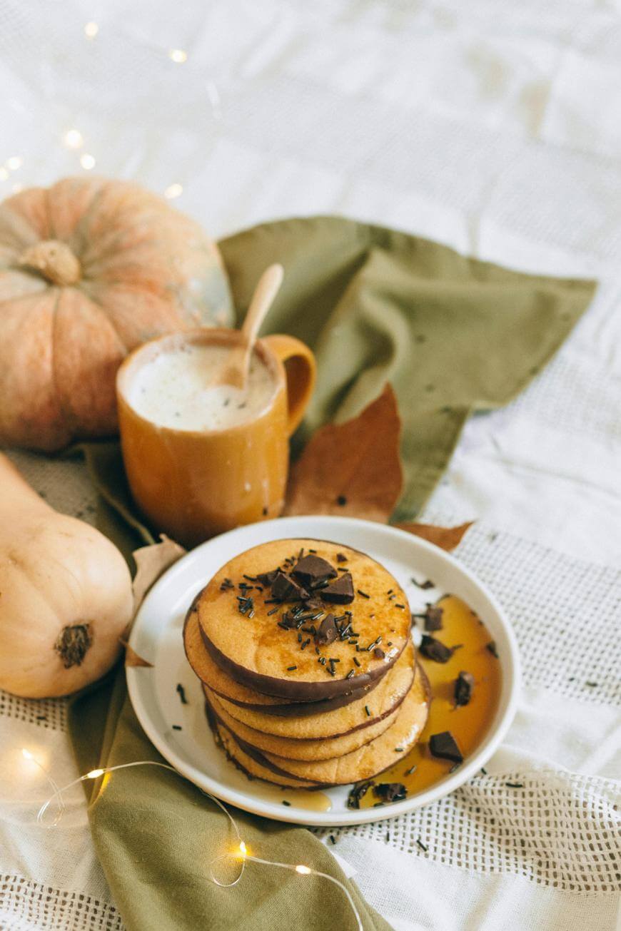 A stack of pancakes topped with chocolate shavings and syrup sits on a white plate next to a yellow mug of frothy drink, surrounded by pumpkins and autumn leaves on a green cloth. Warm fairy lights add a cozy ambiance.