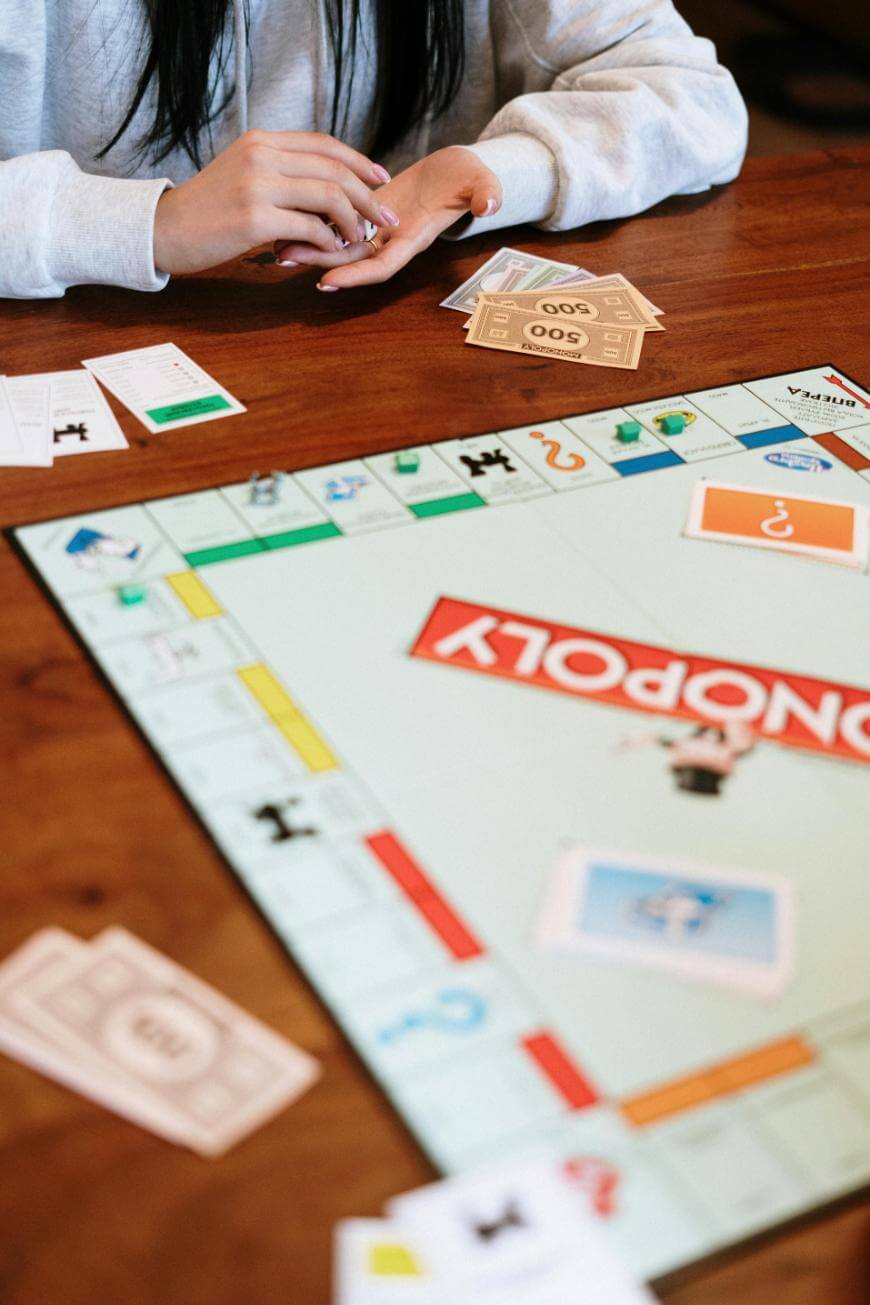 Person playing Monopoly, holding game money and property cards at a wooden table. The board and various cards and bills are spread out in view. Only the persons hands and torso are visible.