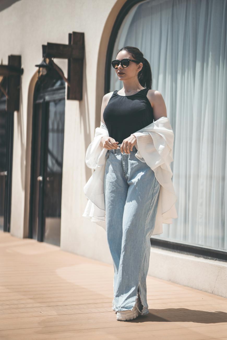 A woman wearing sunglasses, a black sleeveless top, light blue jeans, and a white jacket draped over her shoulders walks on a sunlit terrace in front of large windows.