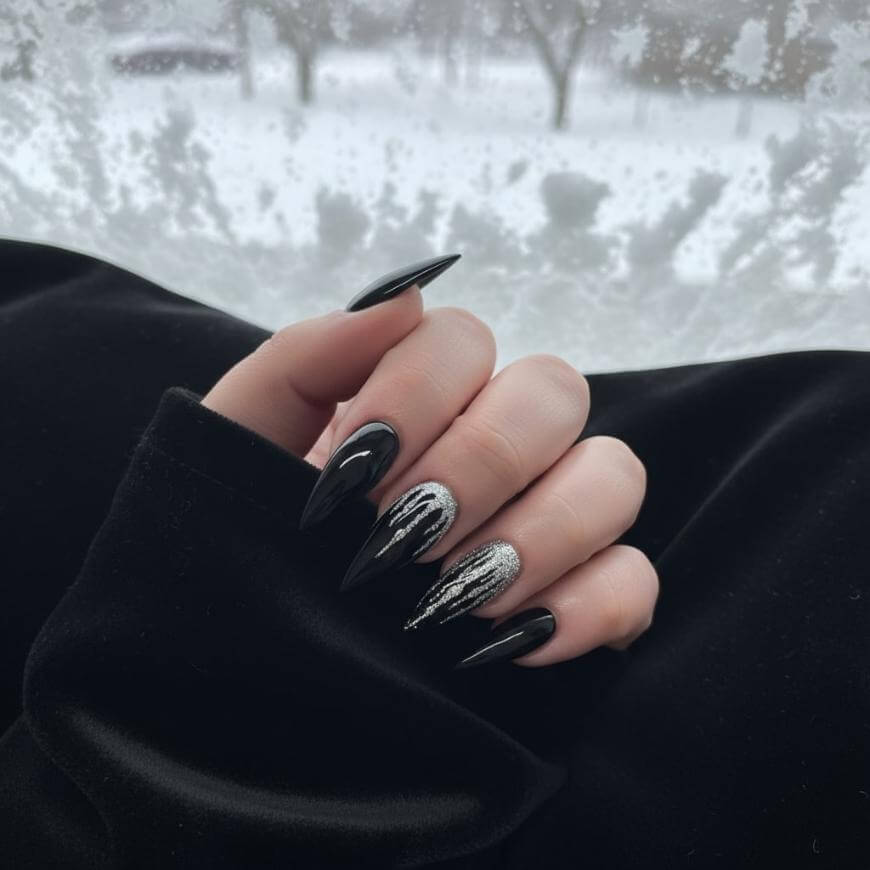 A hand with long, pointed black nails, featuring silver glitter accents on two fingers, is shown against a snowy outdoor background. The person is wearing a black sleeve.