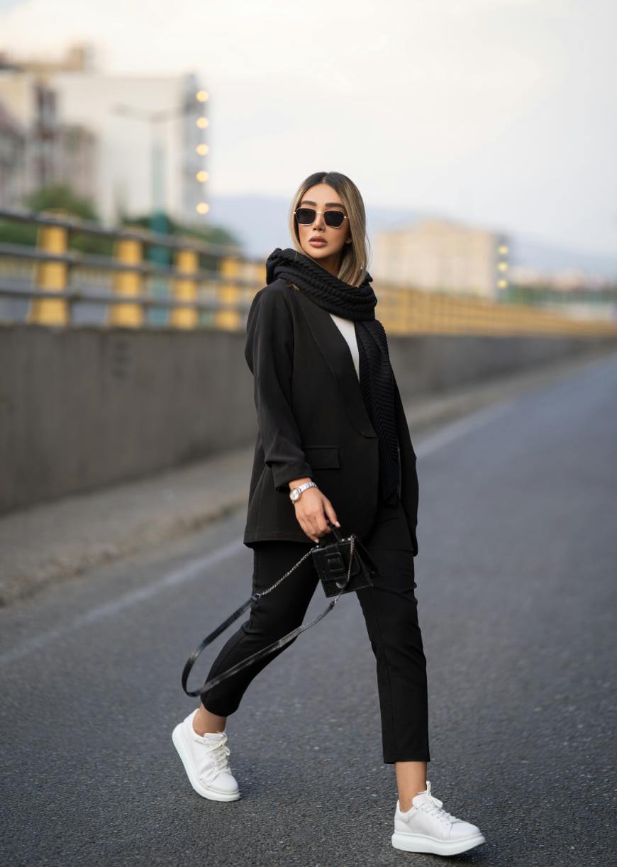 A stylish woman wearing a black suit, scarf, sunglasses, and white sneakers walks confidently on an empty street, holding a small black bag. The background shows an urban setting with buildings and a yellow railing.