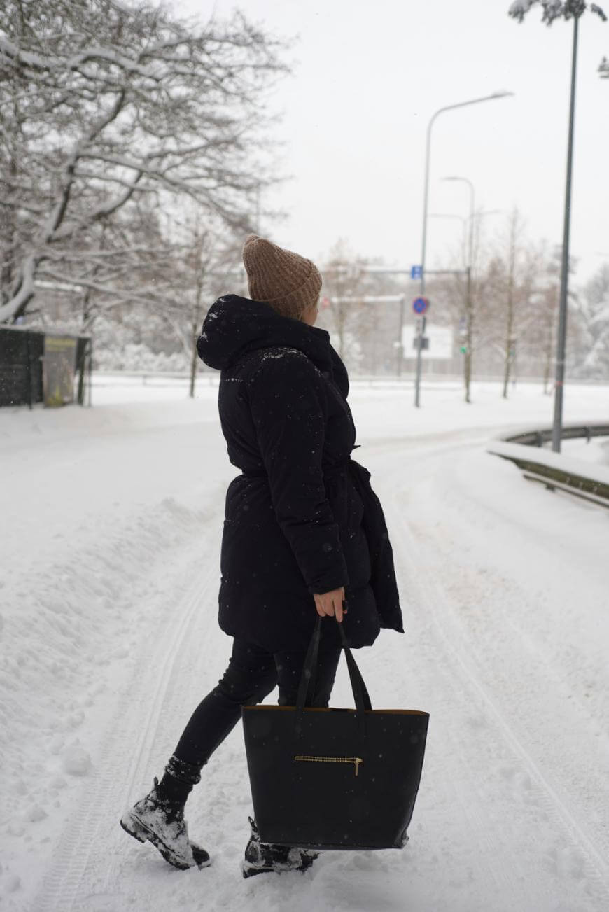 A person wearing a brown knit hat, black winter coat, and boots walks on a snowy street while carrying a large black tote bag. Snow covers the ground, trees, and surroundings.