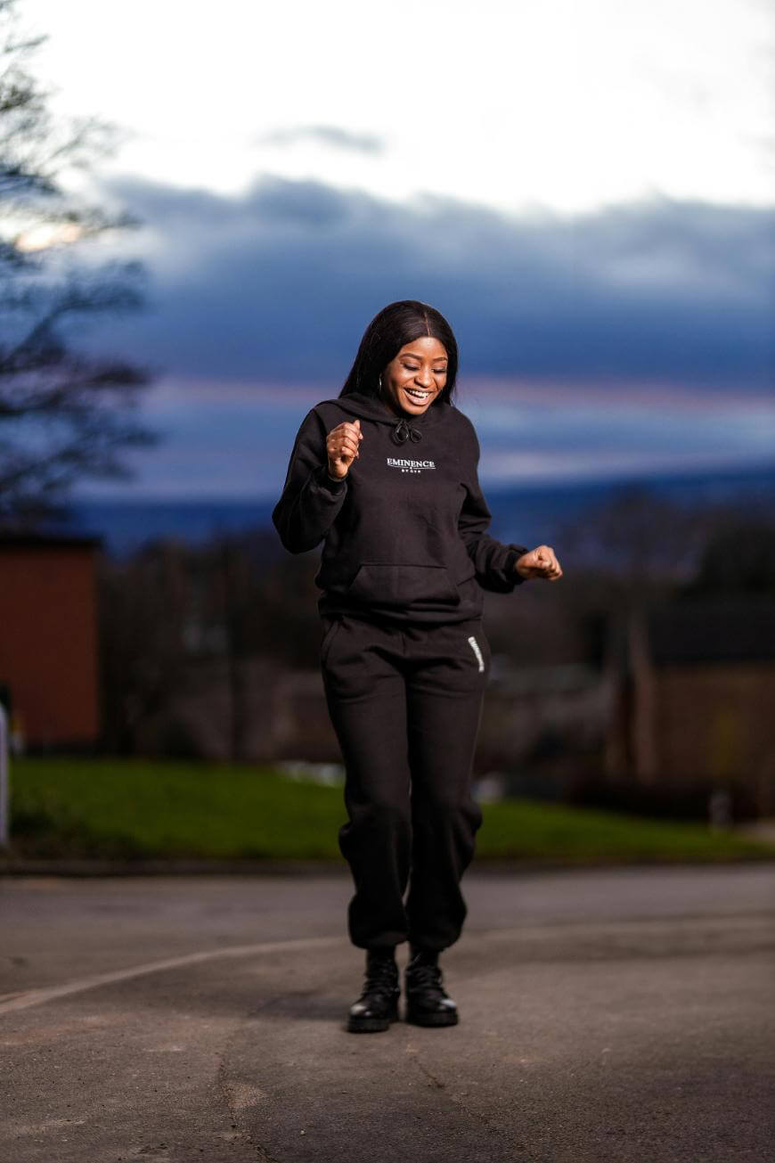 A woman in a black hoodie and sweatpants is smiling and walking outdoors on a paved surface, with houses and a sunset sky in the background.