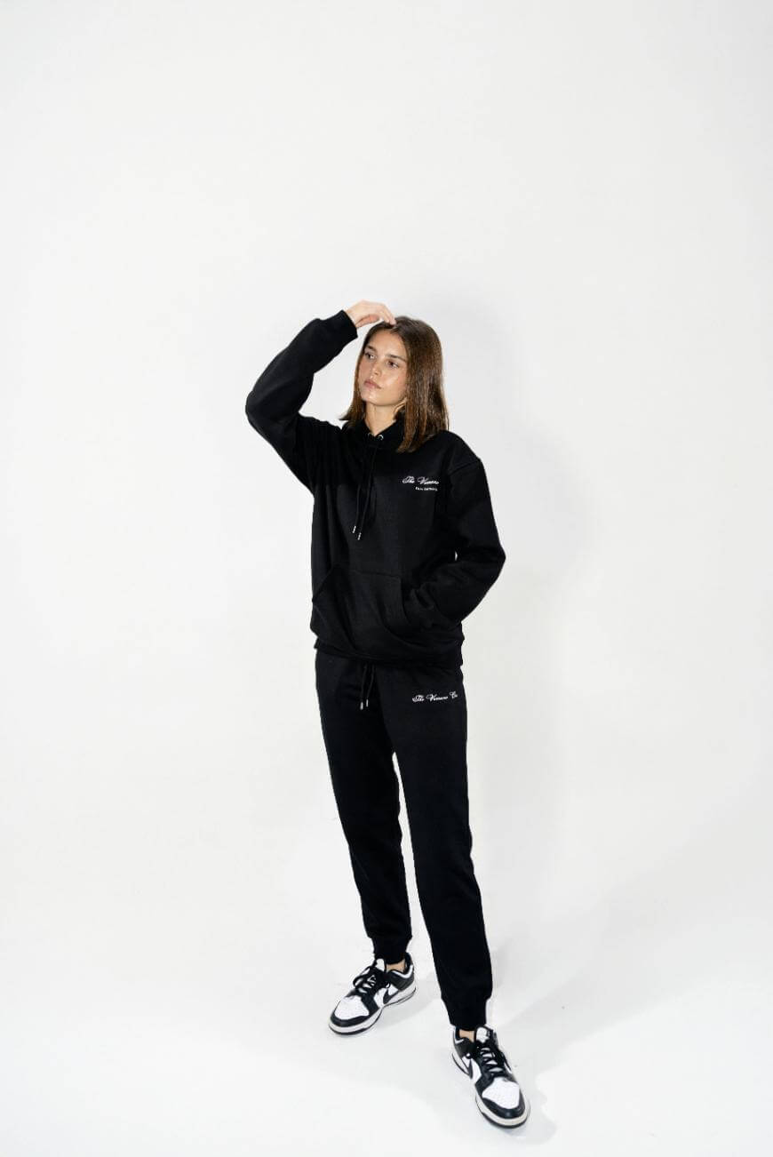 A person stands against a white background wearing a black hoodie, black sweatpants, and black-and-white sneakers, with one hand resting on their head and the other by their side.