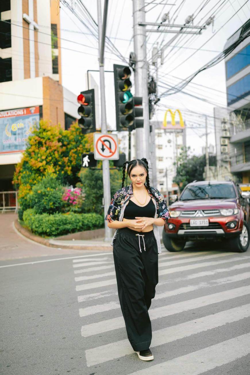 A woman in black pants and a patterned top walks across a city crosswalk. Traffic lights, a red SUV, and a McDonalds sign are visible in the background.