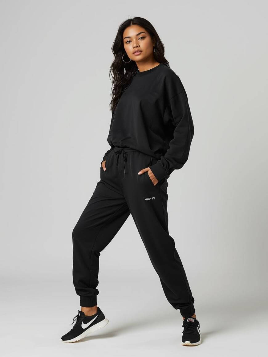 A woman poses confidently in a black sweatshirt and matching joggers with WAIT? text, and black Nike sneakers. She stands against a plain light grey background with hands in her pockets.