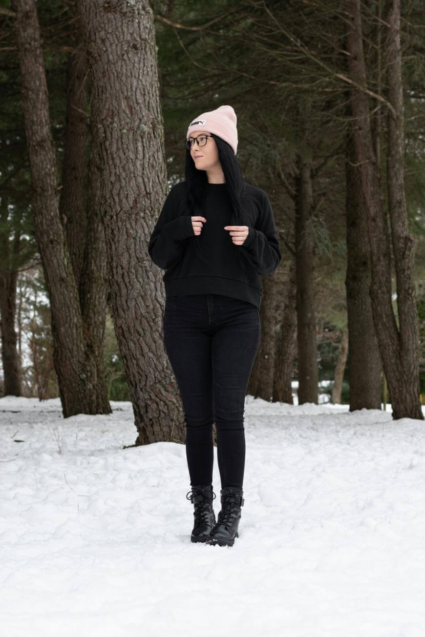 A person wearing a light pink beanie, black glasses, a black sweater, black pants, and black boots stands on snow in a pine forest, looking to the side.