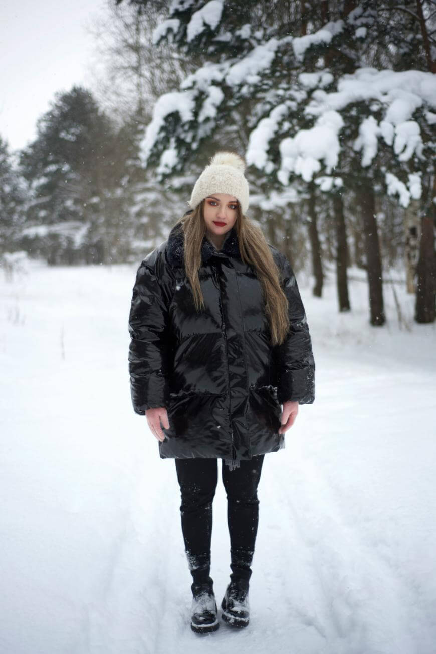A woman stands in a snowy forest wearing a white knit hat, a shiny black puffer jacket, black pants, and black boots. Snow covers the ground and trees, and she looks directly at the camera.