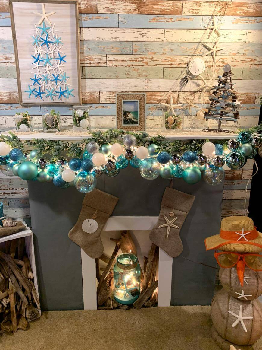 Coastal-themed Christmas mantel with teal, blue, and silver ornaments, starfish decor, stockings, a small tree, and nautical accents, featuring driftwood and a snowman made of stacked beach items.