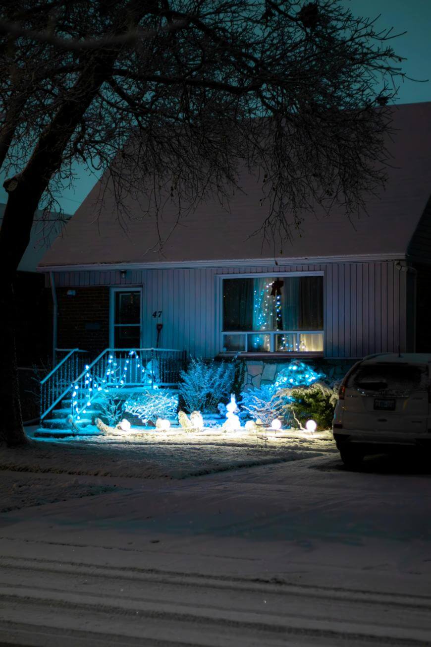 A house with blue and white holiday lights decorating the porch and bushes, a snowman decoration in the yard, snow on the ground and roof, a tree in front, and a car parked in the driveway at night.