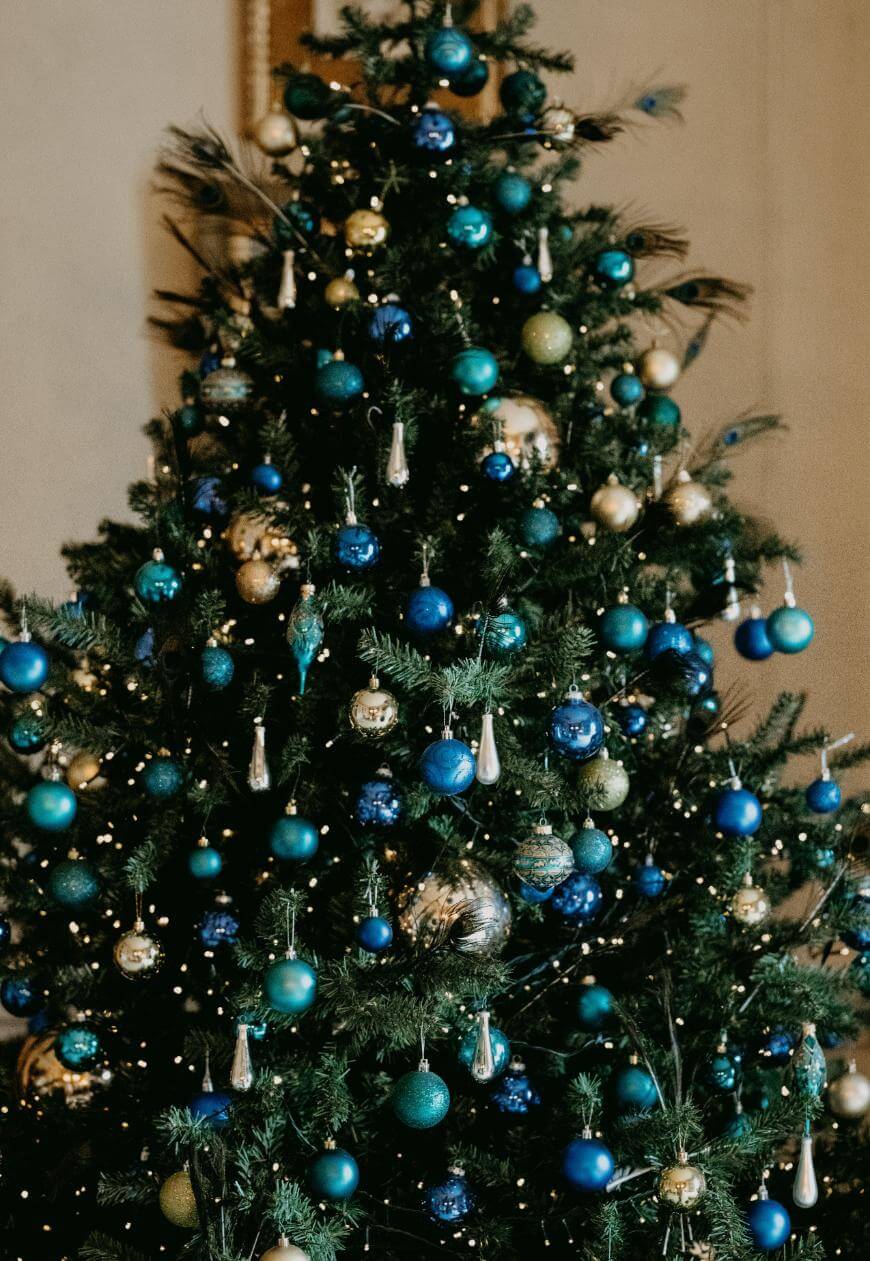 A decorated Christmas tree with green, blue, and gold ornaments and lights, set indoors against a neutral background.