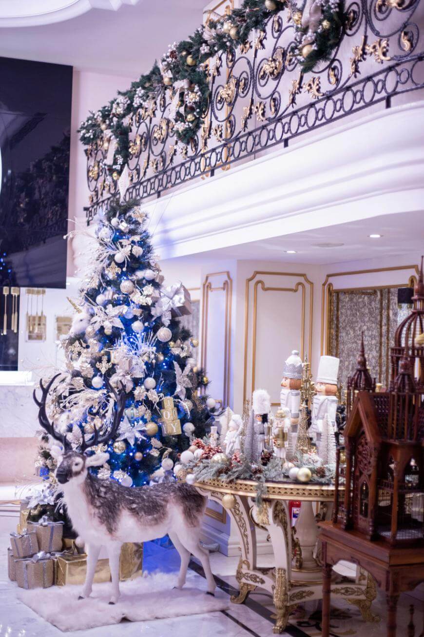 Luxurious Christmas decor in an elegant room featuring a decorated tree, wrapped gifts, a realistic reindeer figure, festive garlands on a balcony, and a table with holiday ornaments and nutcrackers.