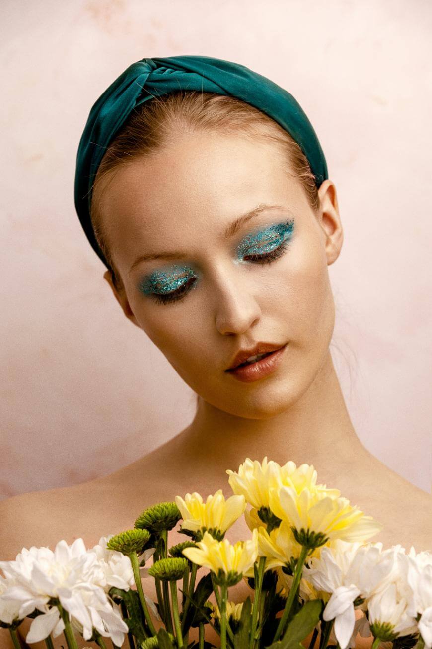 A woman with blue glitter eyeshadow and a green headband looks down, surrounded by yellow and white flowers against a soft pink background.
