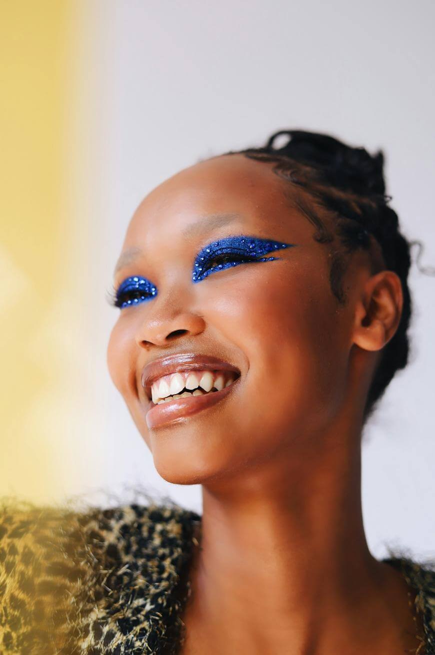 A smiling person with braided hair, wearing bold blue glitter eye makeup and a textured, leopard-patterned outfit, looks off-camera against a light background.