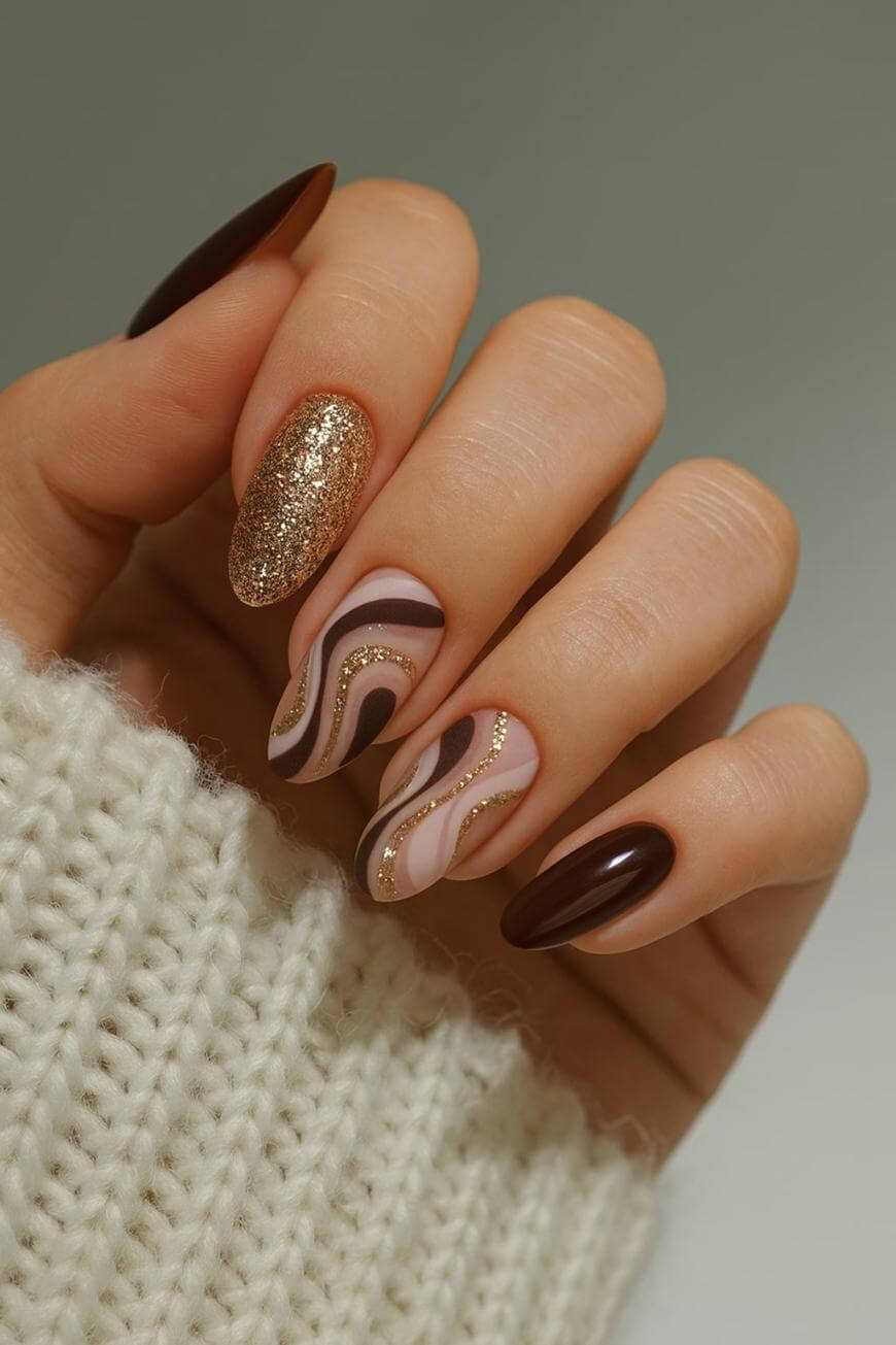 A hand with long almond-shaped nails features a brown manicure: one nail is glittery gold, two have wavy brown and gold patterns, and two are solid brown. The person wears a cream knitted sweater.