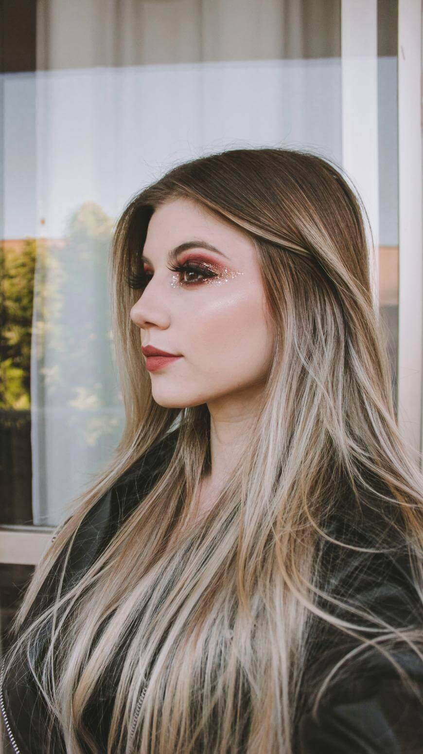 A woman with long, light brown hair and glittery red eye makeup is shown in profile, standing in front of a window with a curtain and greenery visible outside. She is wearing a black jacket.