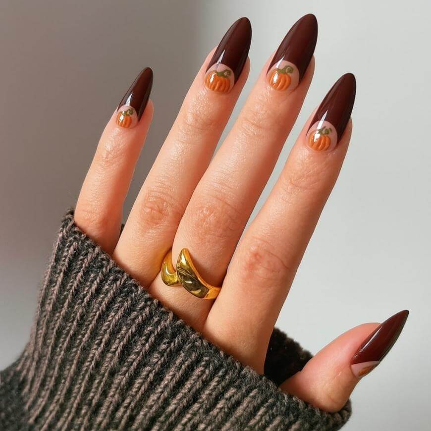 A hand with long, almond-shaped nails painted dark brown, featuring small orange pumpkin nail art near the cuticles. The person wears a gold ring and a ribbed, dark-colored sweater.
