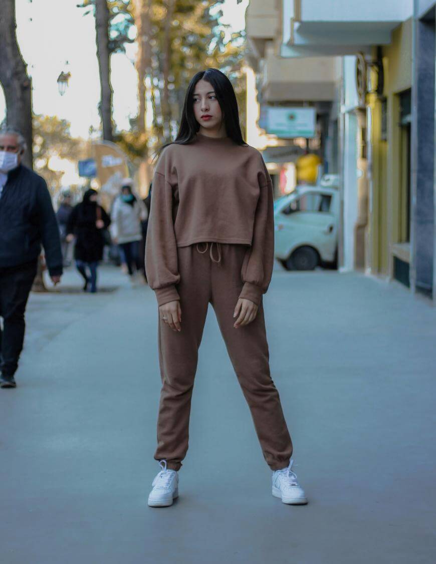 A young woman with long dark hair stands confidently on a city sidewalk, wearing a brown matching sweatshirt and joggers set with white sneakers. People and trees are visible in the background.