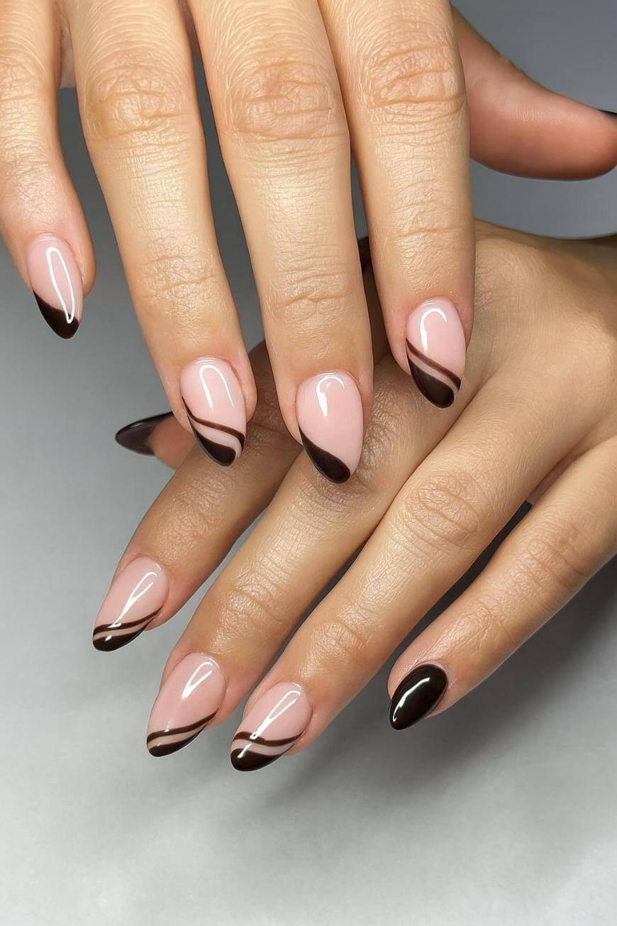 A close-up of hands with almond-shaped nails featuring a nude base and dark brown tips, accented with a thin brown diagonal stripe near the tips. The background is plain and light-colored.